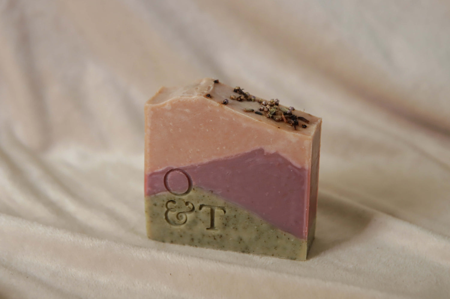 Heather soap bar out of packaging, design of three colours replicating Scottish hills, a green grassy base, a gentle purple middle of heather and a pink top layer of a pink sunset sky, topped with lavender and heather and stamped with the O&T logo